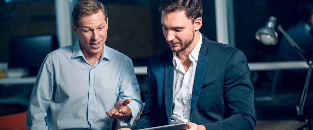 Two office workers with tablet in office late at night. Young businessmen talking while viewing presentation on digital touchscreen tablet.